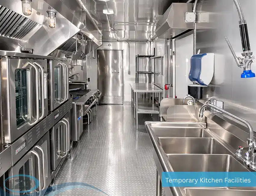Interior of a temporary commercial kitchen trailer with stainless steel prep tables, triple-compartment sink, commercial ovens, cooking equipment, and ventilation system.